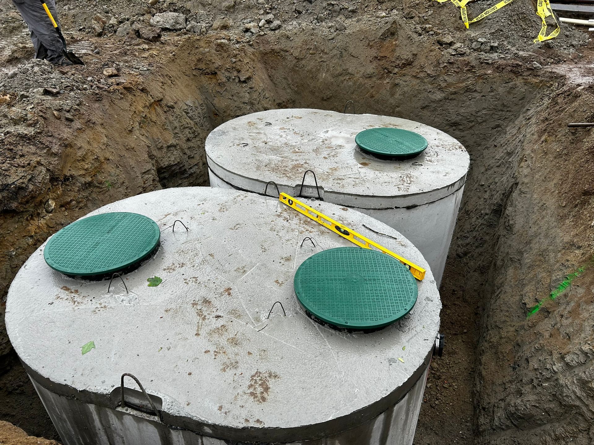 Two concrete septic tanks in a dug pit, each with green access lids. A level and a shovel are visible.