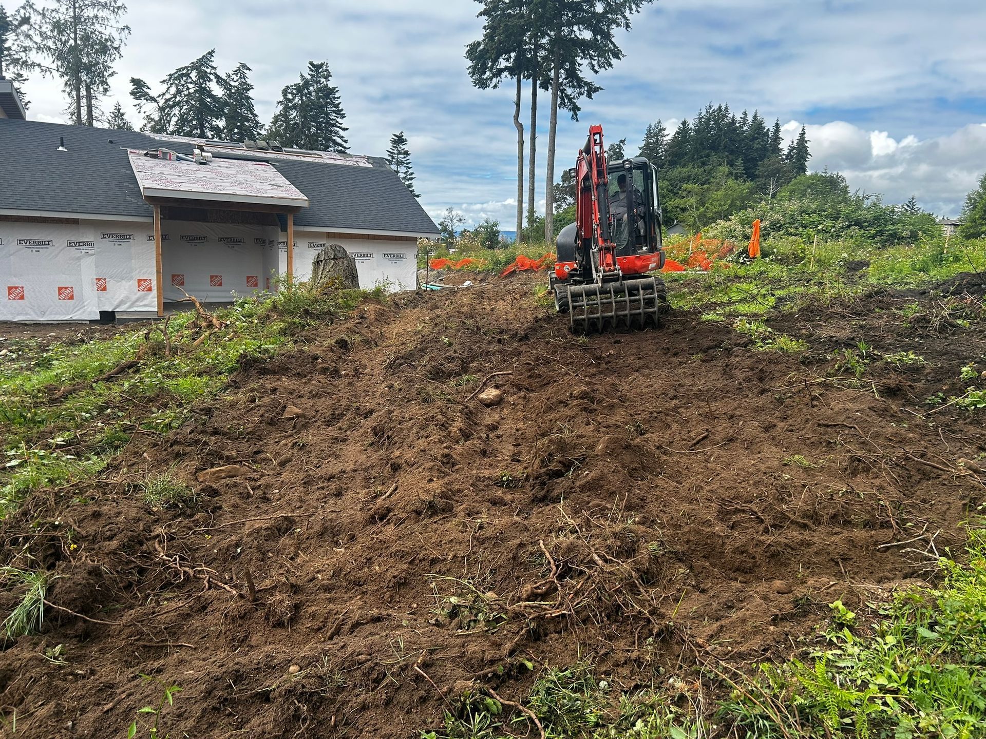An excavator working on a dirt mound in front of a partially built house on a cloudy day.