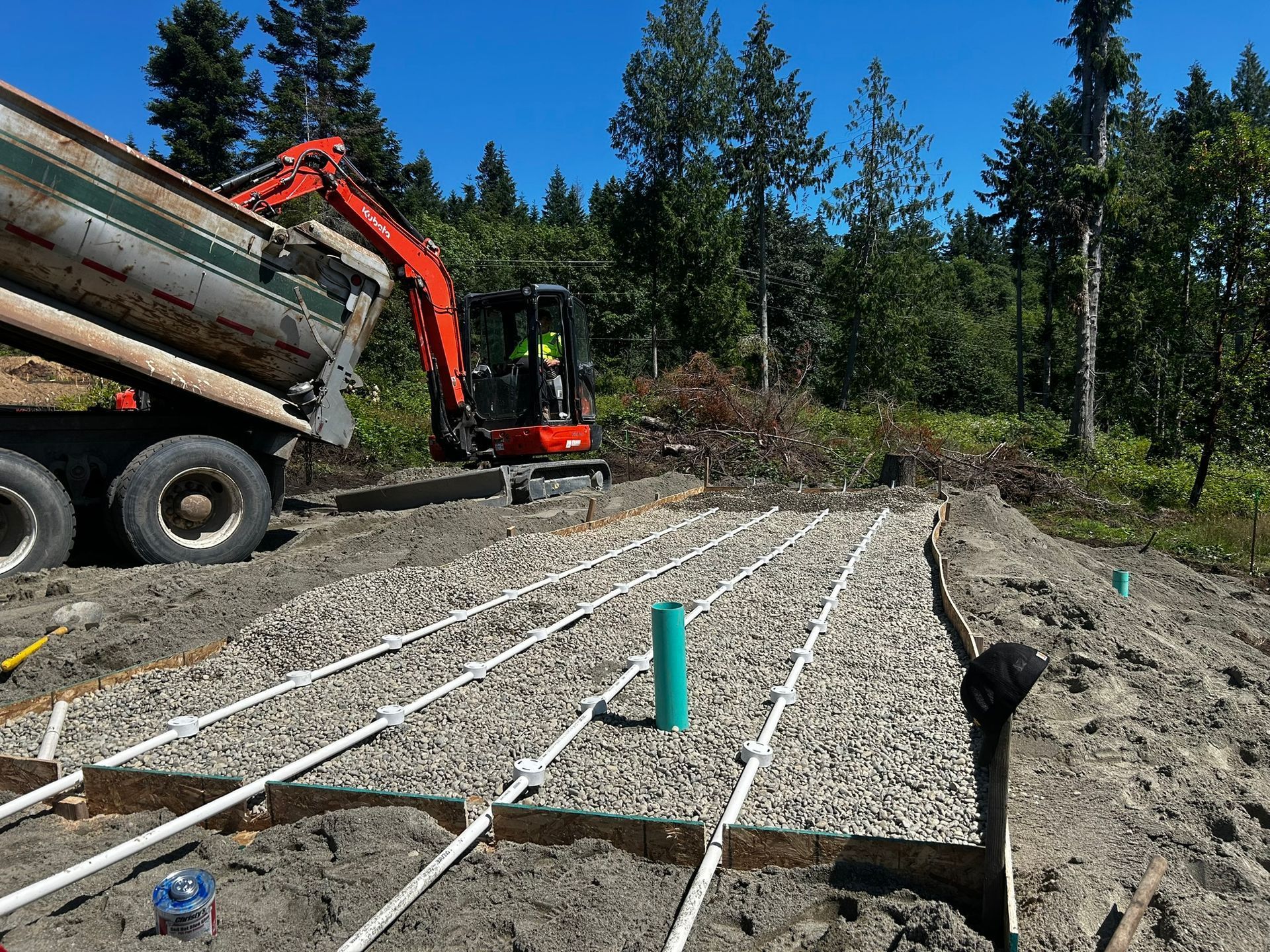 Construction site with a truck dumping gravel onto a lined trench. An excavator and trees are in the background.