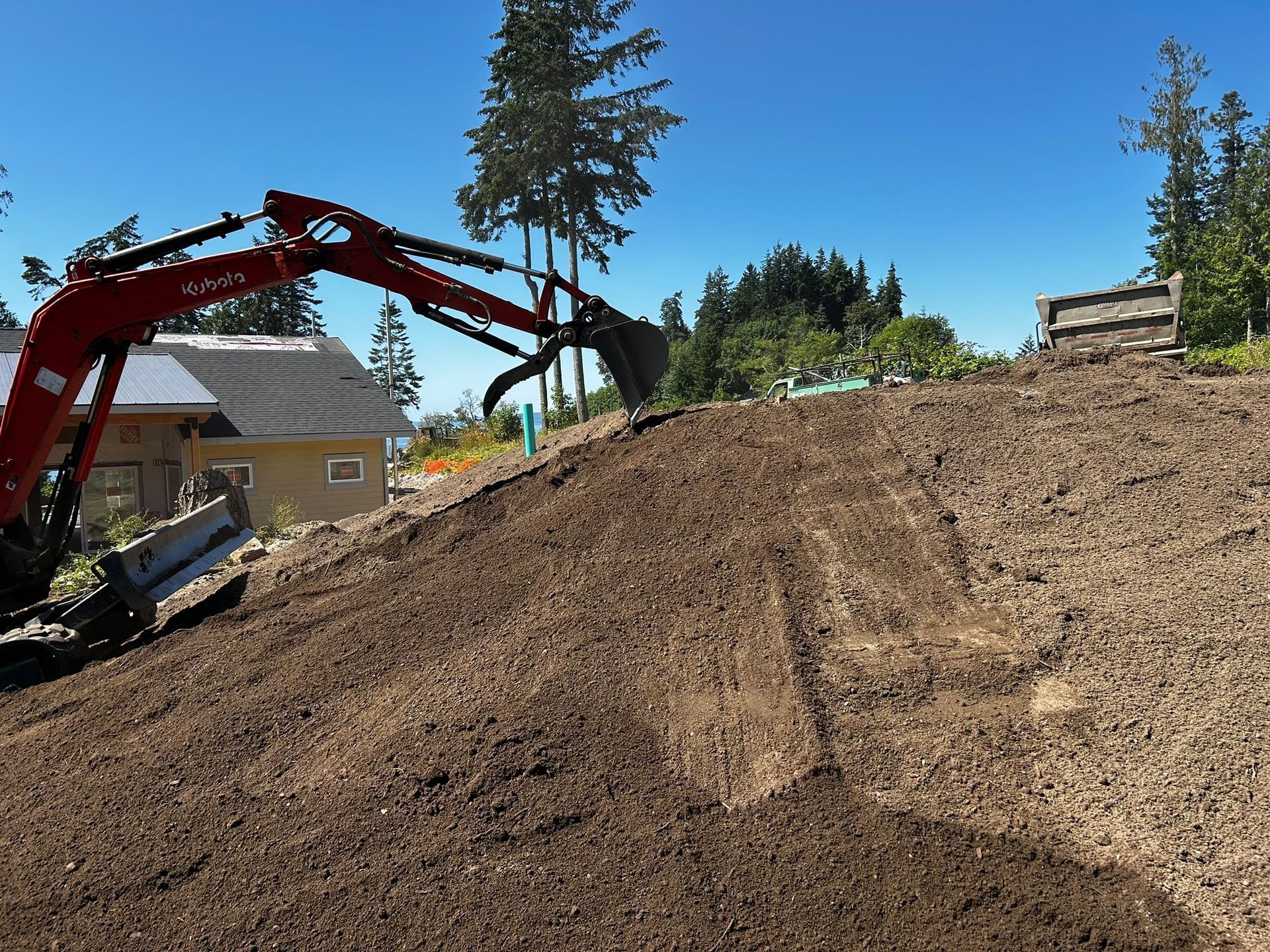 Red excavator leveling dirt on a hillside near a house and trees under a blue sky.