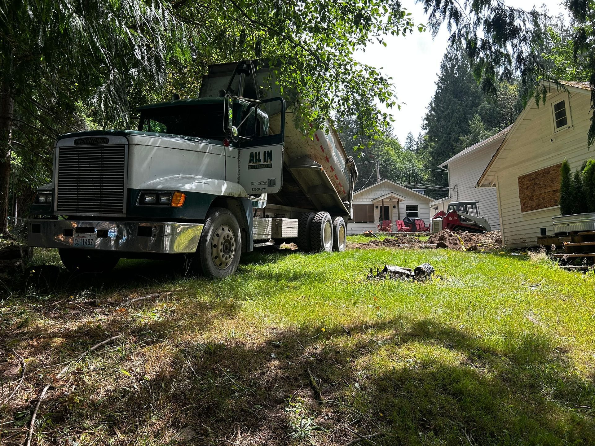 Dump truck parked on a grassy area near a house, with construction debris and another vehicle visible in the background.
