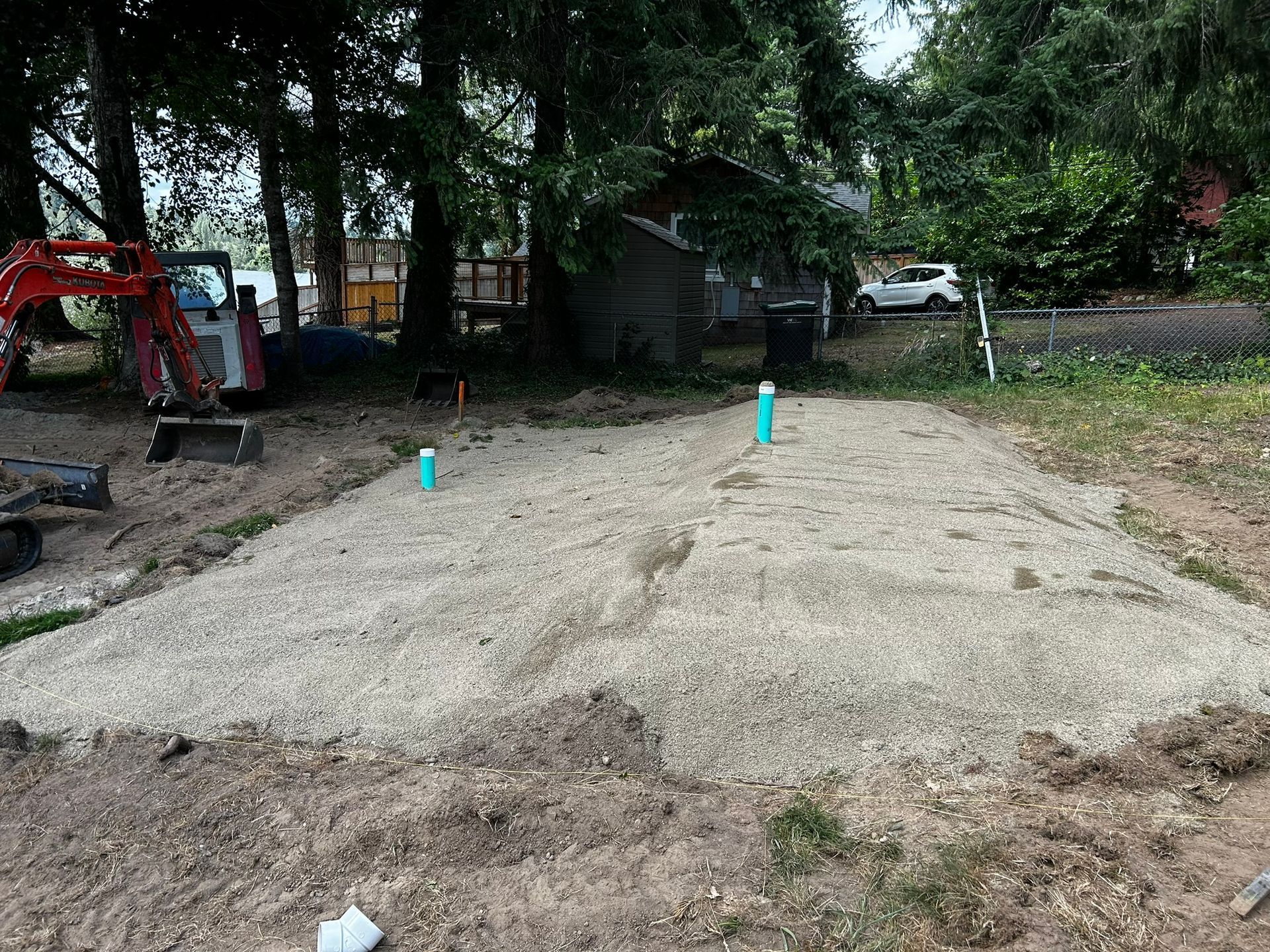 A gravel-filled area with blue plumbing pipes; an excavator sits nearby, trees in the background.