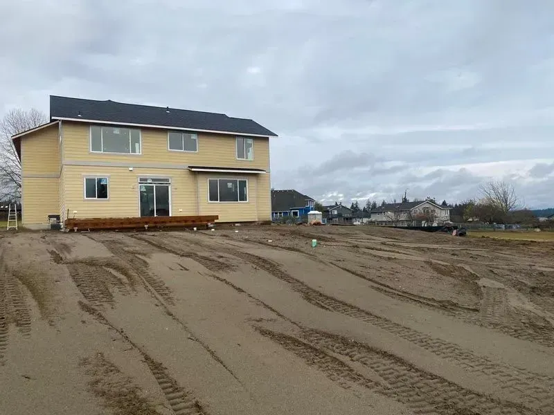 Two-story house with unfinished yard. Yellow siding, black roof, sliding glass door, and bare ground with tire tracks.