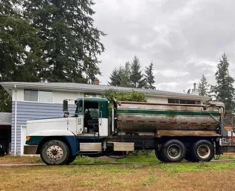 Old fuel truck parked on a grassy lot, in front of a house. White, green, and silver colors. Cloudy day.