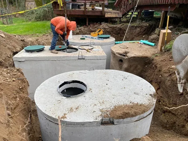 Person installing septic tanks in an excavation. Concrete tanks with open lids and tools visible outdoors.