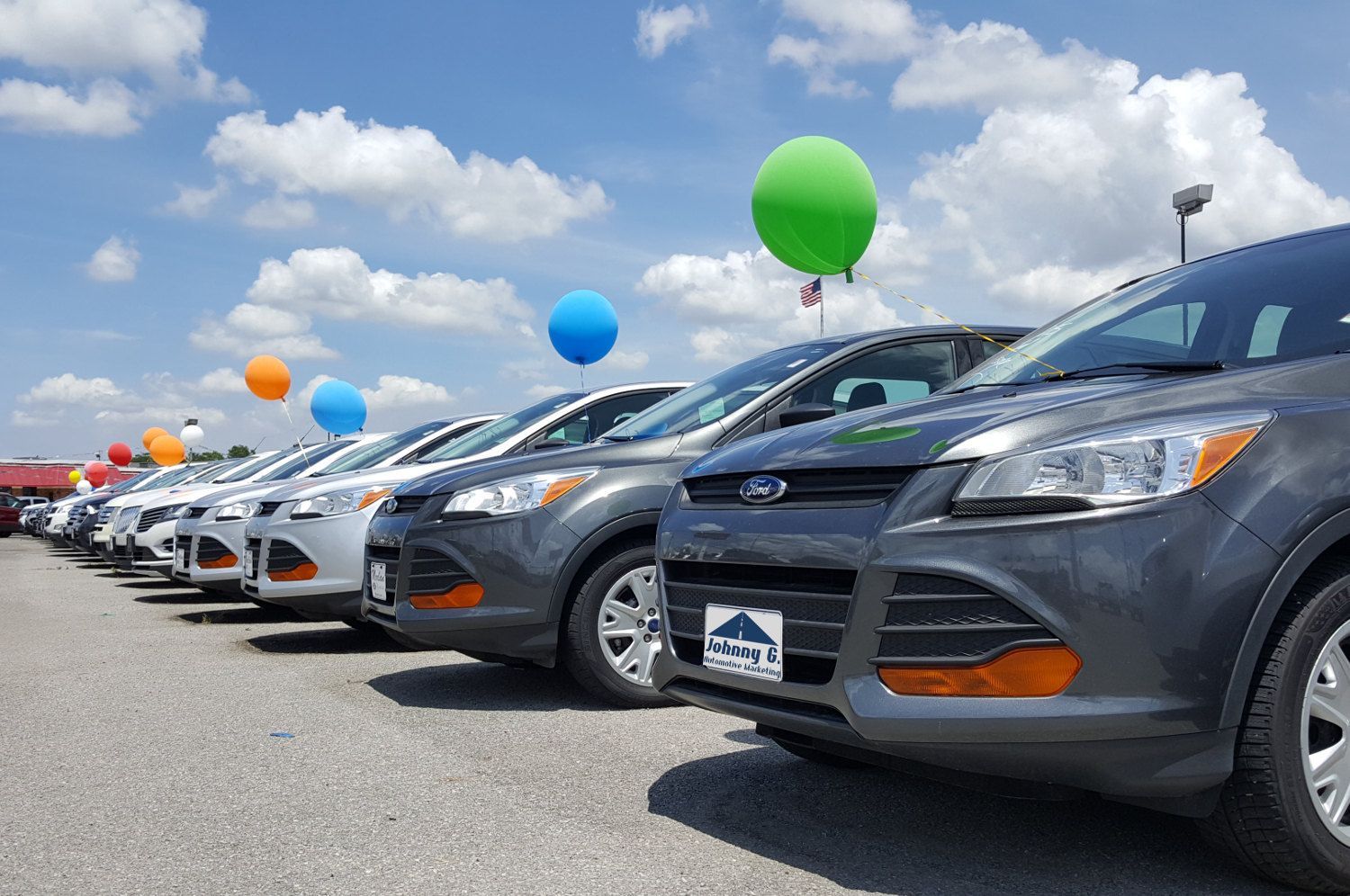 Cars lined up at a dealership lot with colorful balloons under a blue sky