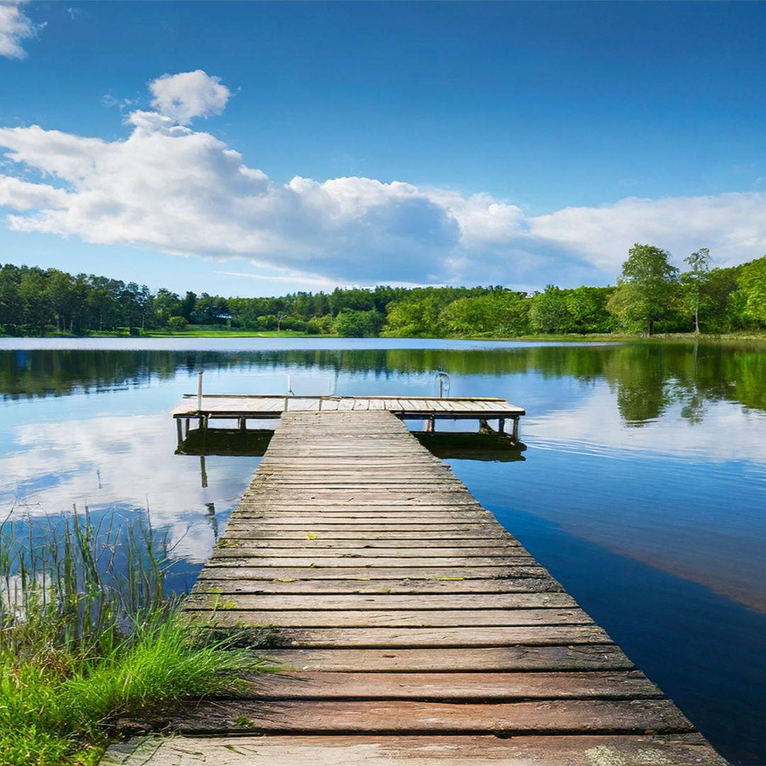 A wooden dock stretches into a calm, blue lake, surrounded by a lush green forest under a sky with light, scattered clouds.