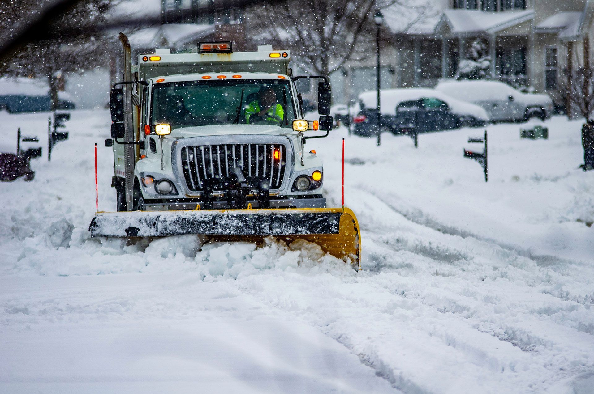 White truck with yellow snow plow blade clearing streets after heavy snowfall in urban area with driver wearing yellow safety vest