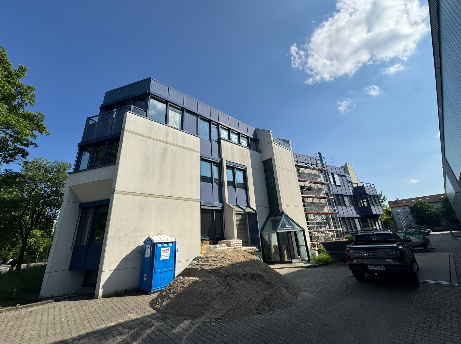 Gebäude mit blauem Dach und schräger Fassade, blaue mobile Toilette, Kieshaufen, geparkte Fahrzeuge, blauer Himmel.