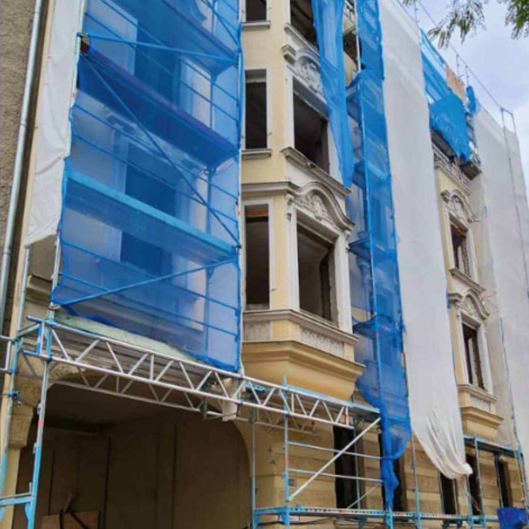 Das Gebäude wird renoviert und ist mit Gerüsten und Sicherheitsnetzen verhüllt. Weiß und blau. Der Gehweg ist durch Absperrungen blockiert.