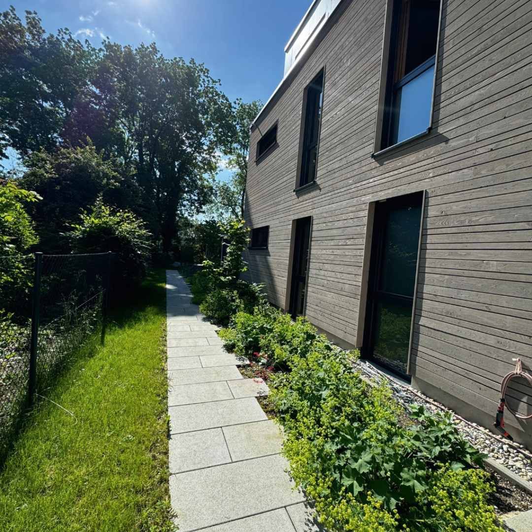 Steinweg neben einem modernen Haus mit Holzverkleidung, Gartenbeet und Bäumen, sonniger Tag.