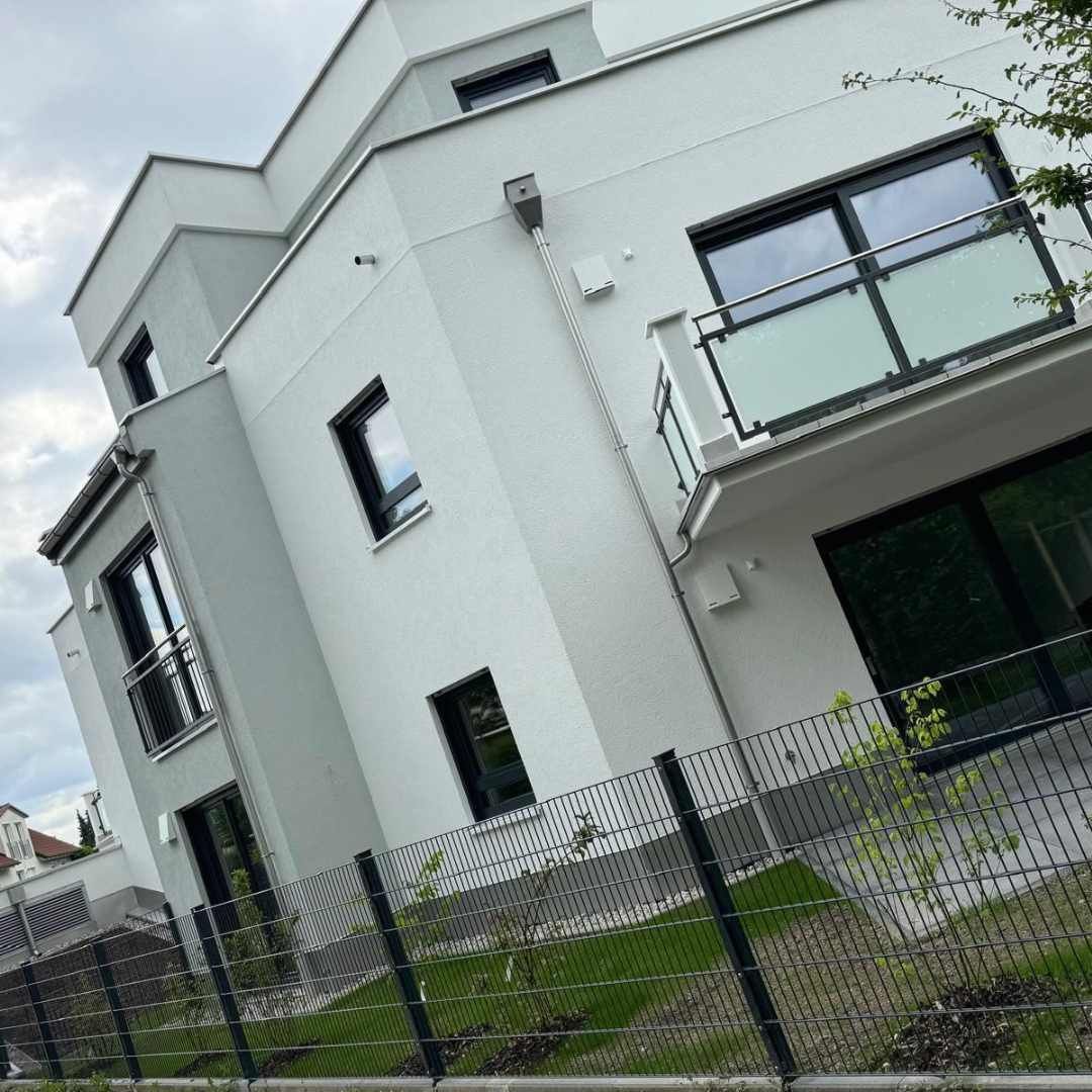 Modernes, weißes, mehrstöckiges Gebäude mit schwarz umrandeten Fenstern und einem kleinen Balkon, hinter einem schwarzen Metallzaun.