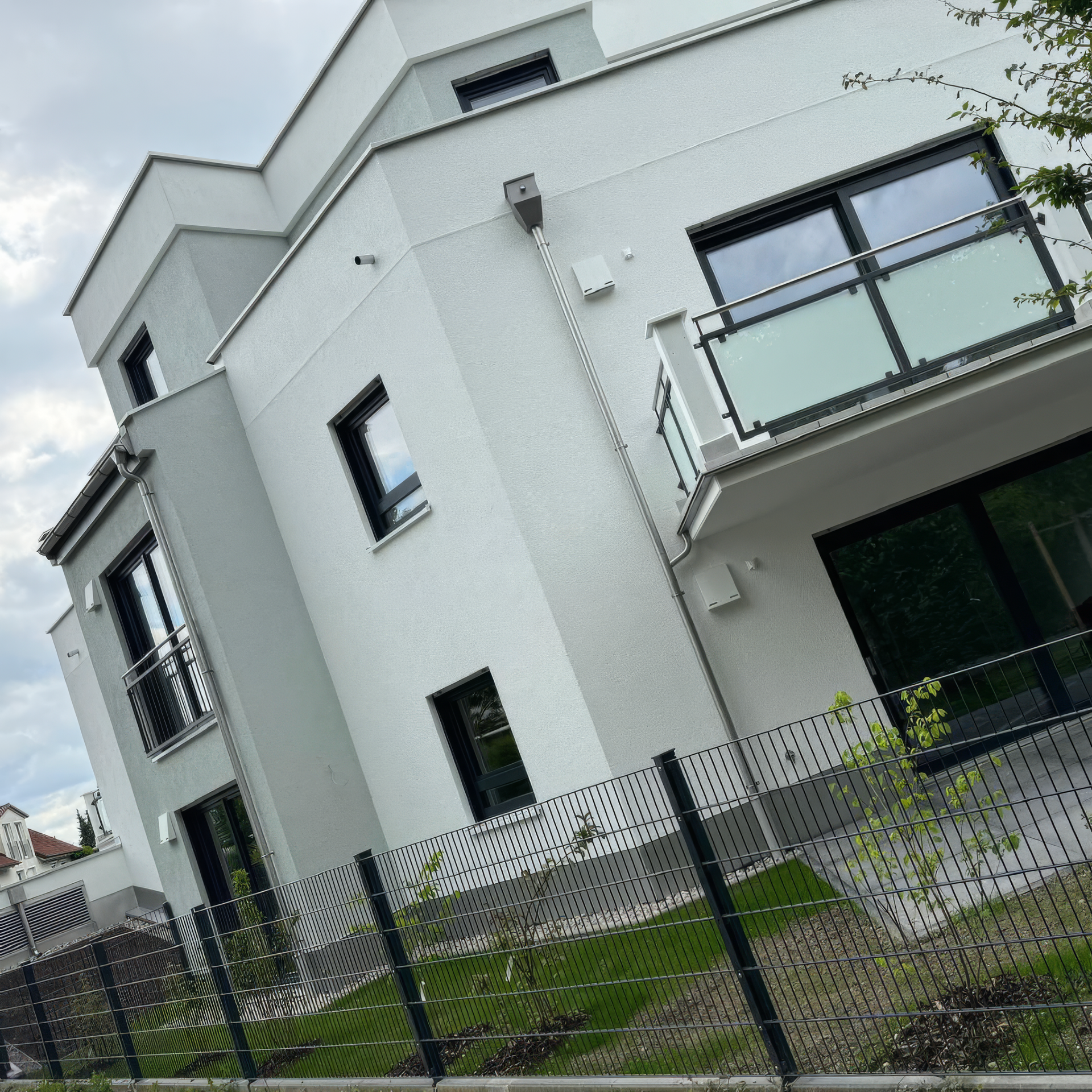 Modernes, weißes, mehrstöckiges Gebäude mit schwarz umrandeten Fenstern und einem kleinen Balkon, hinter einem schwarzen Metallzaun.