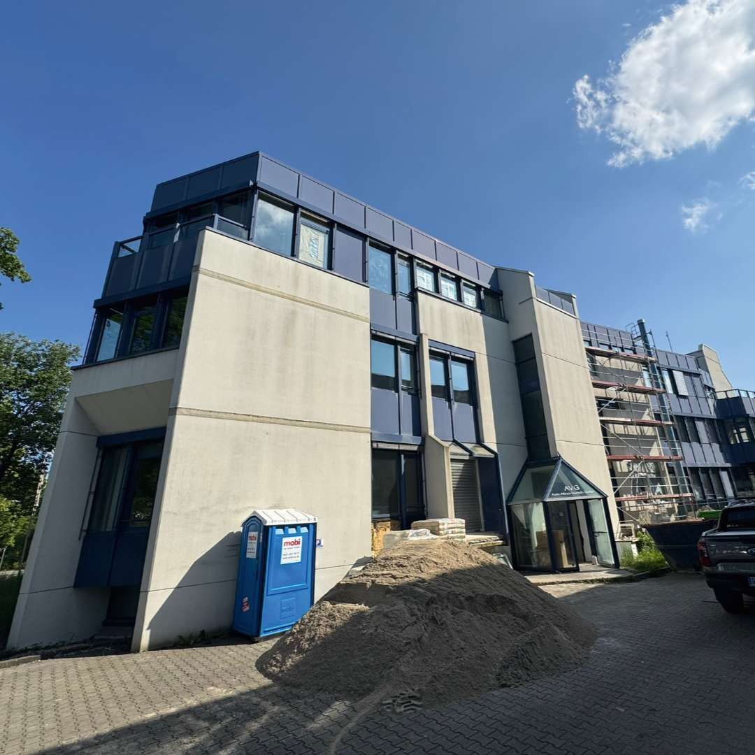 Gebäude mit blauem Dach und schräger Fassade, blaue mobile Toilette, Kieshaufen, geparkte Fahrzeuge, blauer Himmel.