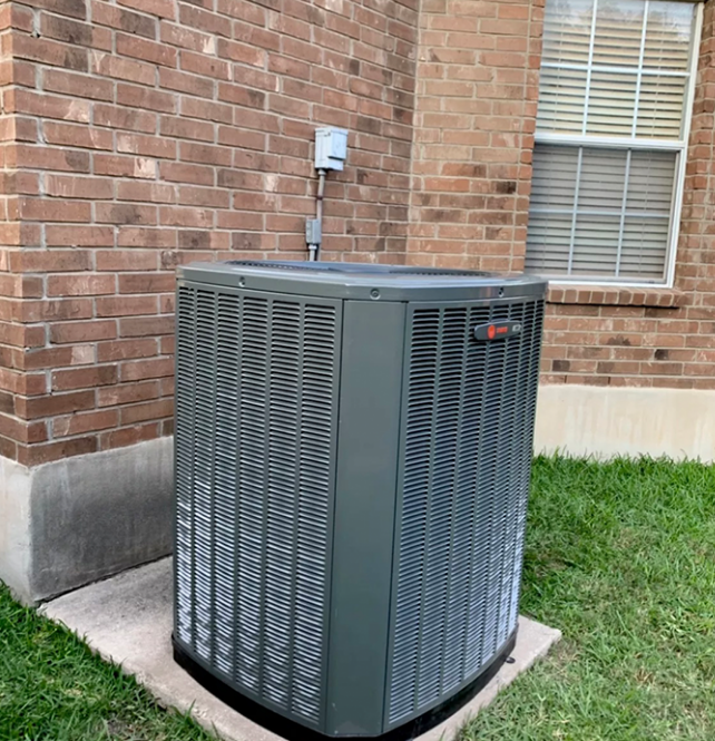 An outdoor air conditioning unit against a brick wall, grass, and window.