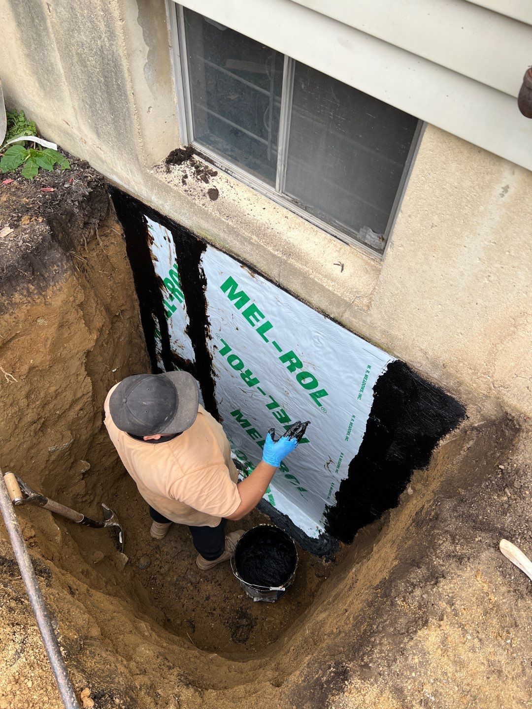 Person applying waterproofing to a basement wall in an excavated area; green and black materials.
