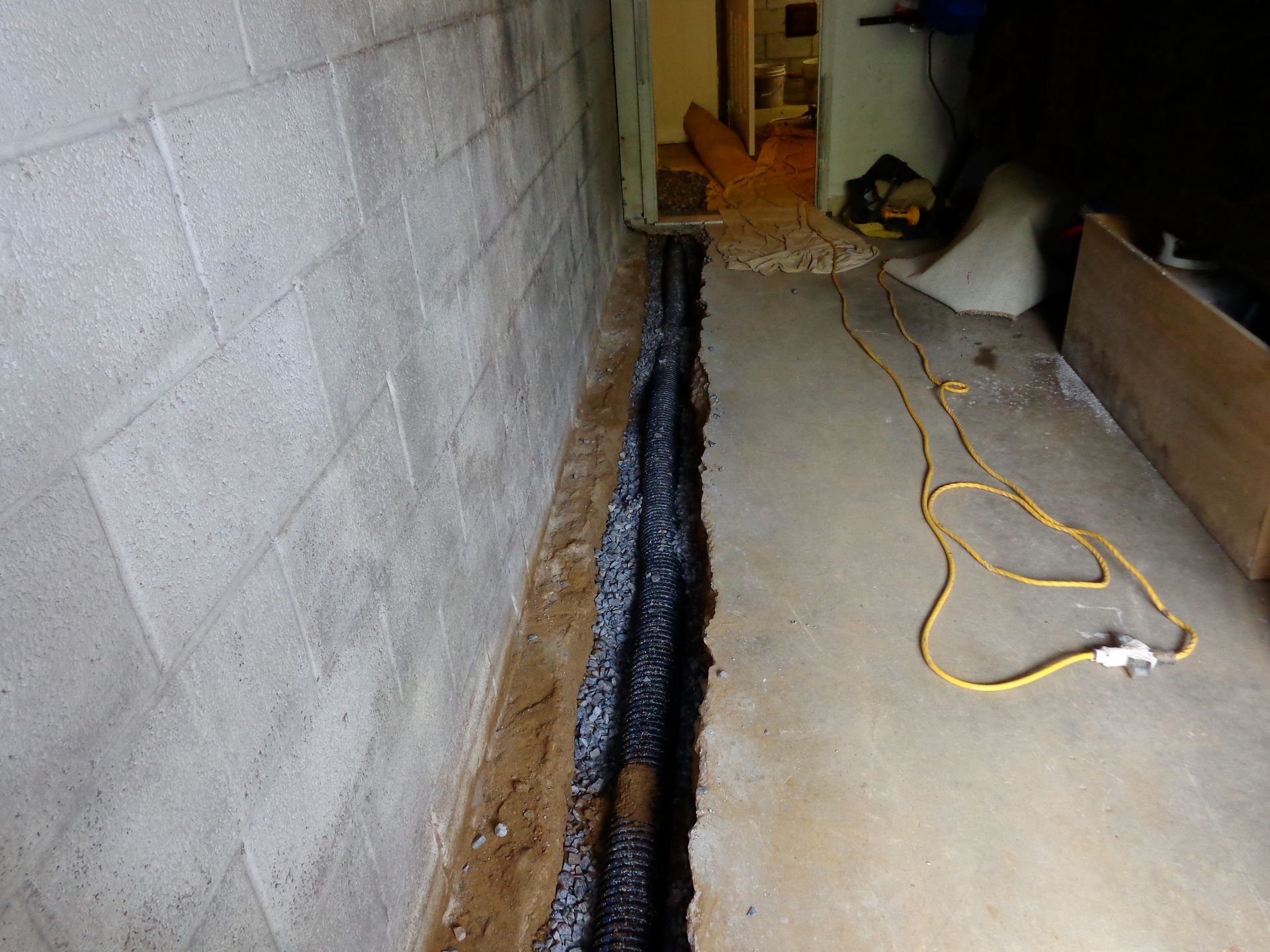 A dark trench dug along a concrete basement wall; black gravel and pipe visible. A yellow extension cord lies nearby.