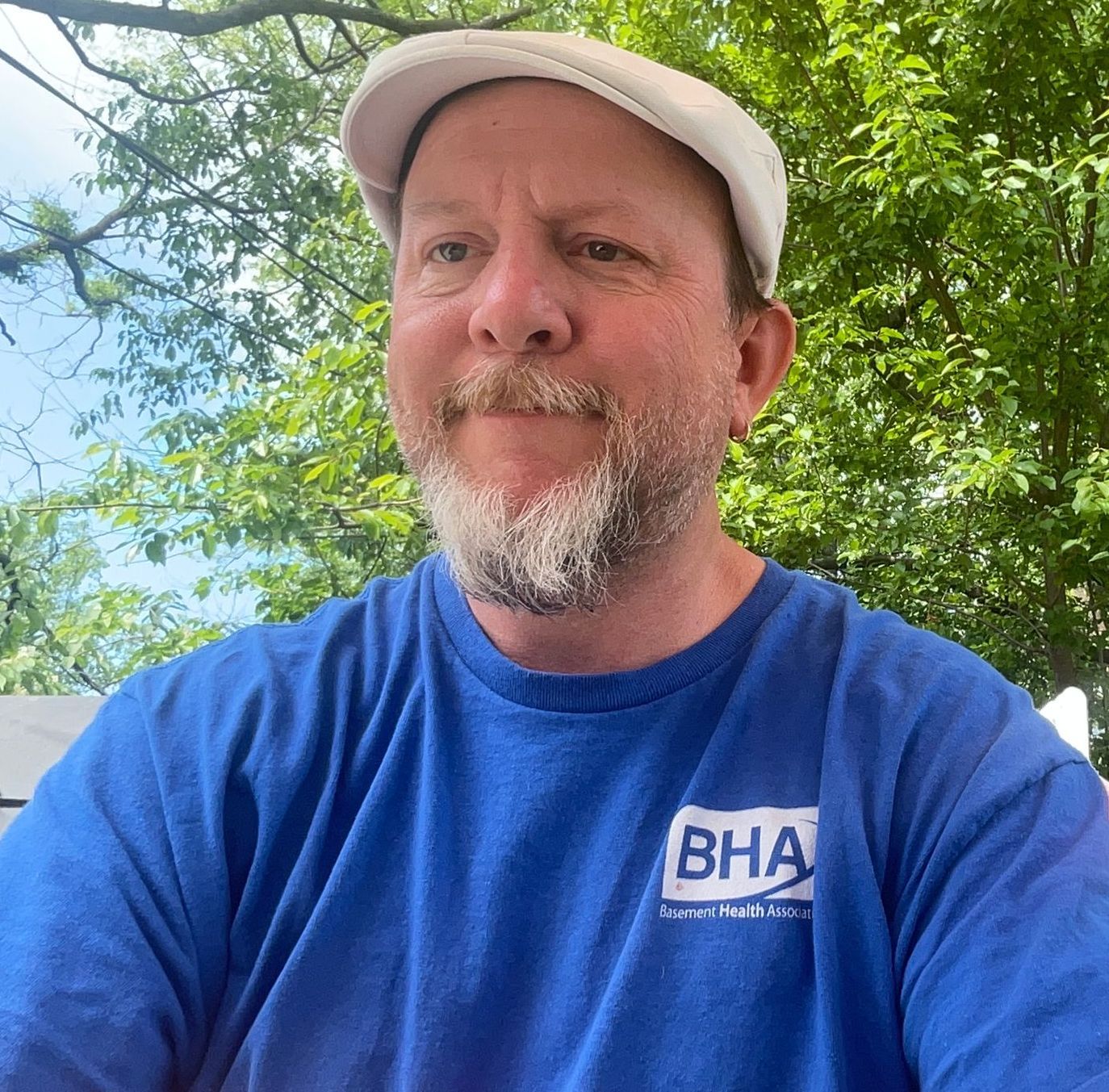 Man with a beard and a white cap smiling outdoors, wearing a blue 