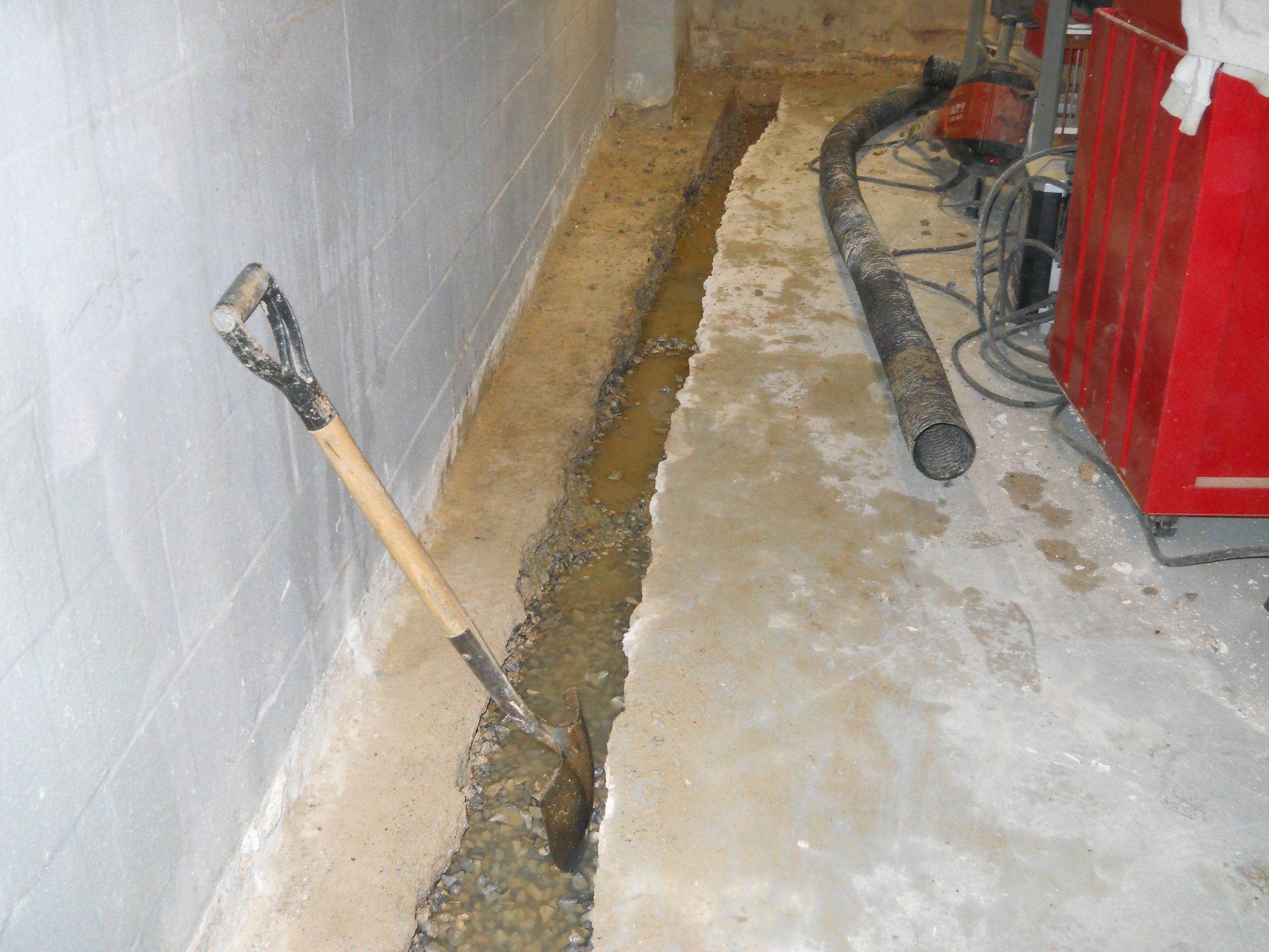 Trench dug in concrete floor along basement wall; shovel rests in trench, black pipe and red object are visible.