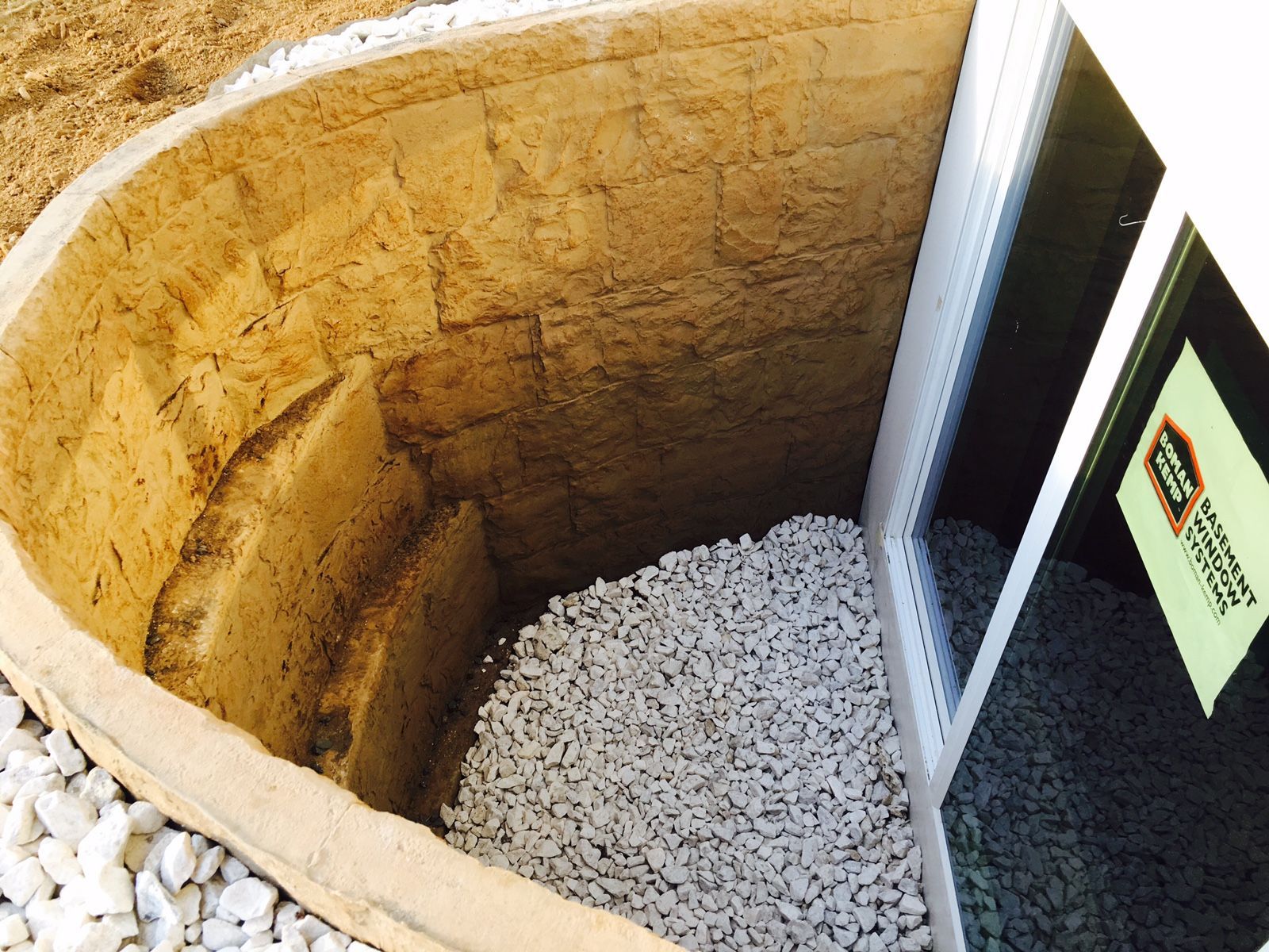Window well with steps, filled with white gravel, next to a basement window. Beige stone walls.