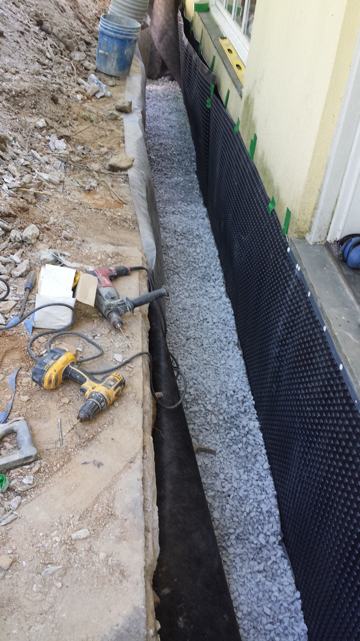 Construction site: trench along a building wall, filled with gravel and wrapped in black drainage membrane.