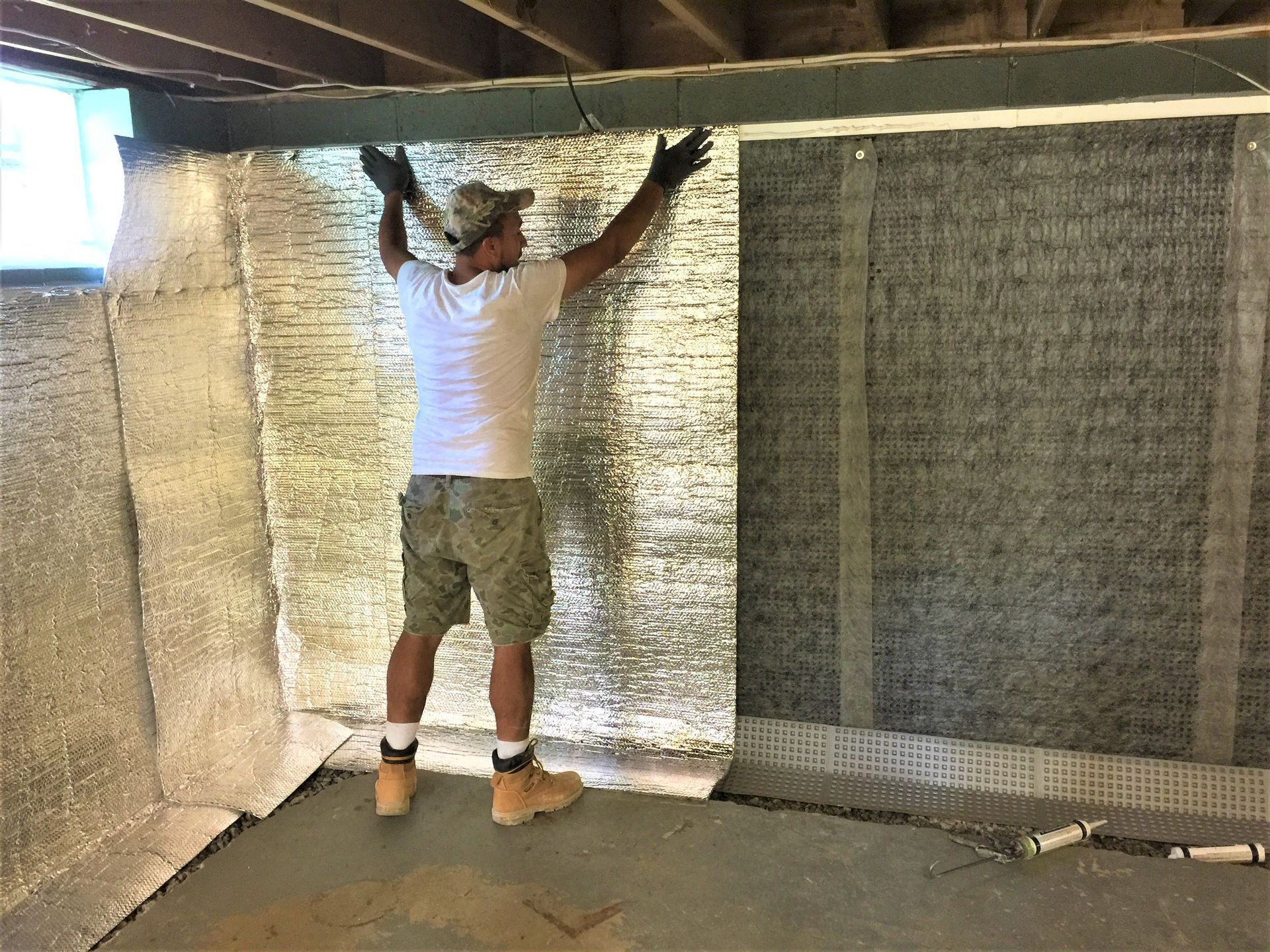 Man installing insulation in a basement. He is standing with arms raised, near silver material and a concrete floor.