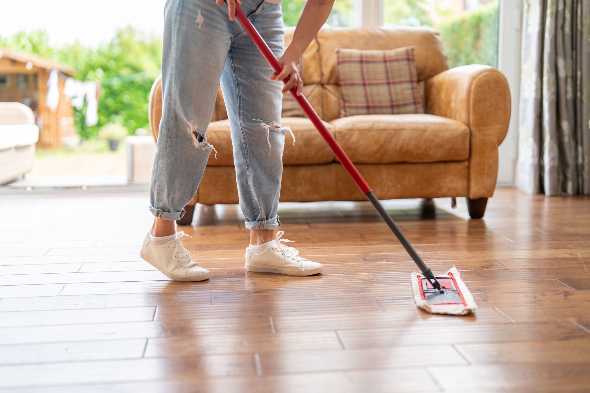 Person mopping a hardwood floor in a living room, wearing jeans and white sneakers.
