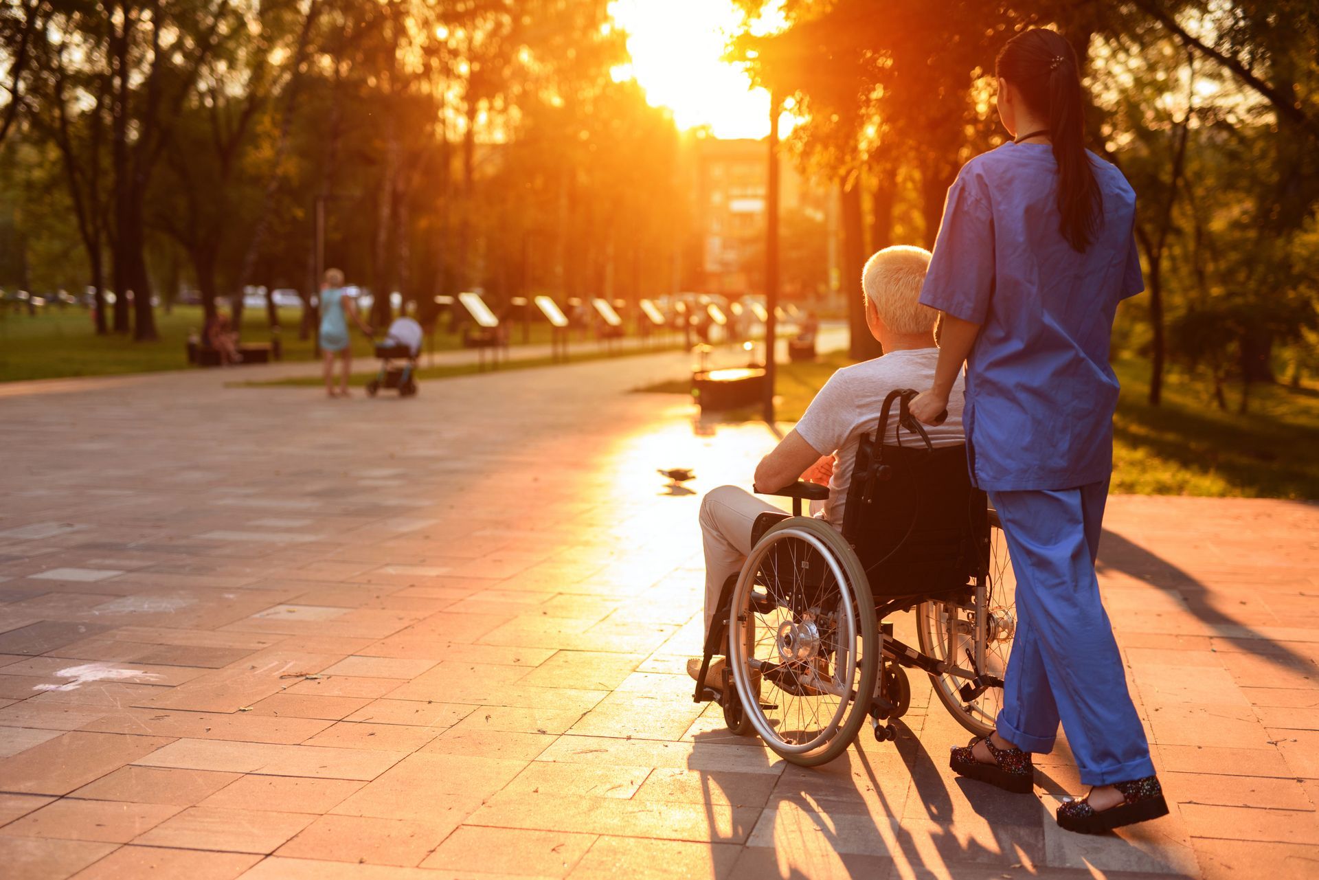 A person in a wheelchair is pushed by a caregiver on a sunny park path.