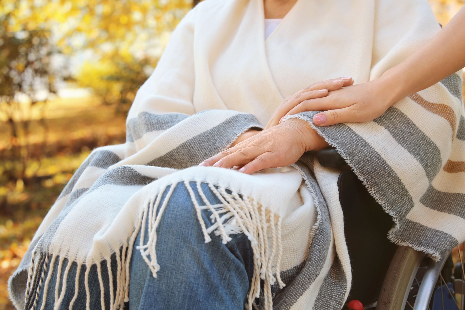 Person in wheelchair with blanket, hand held by another person outdoors.