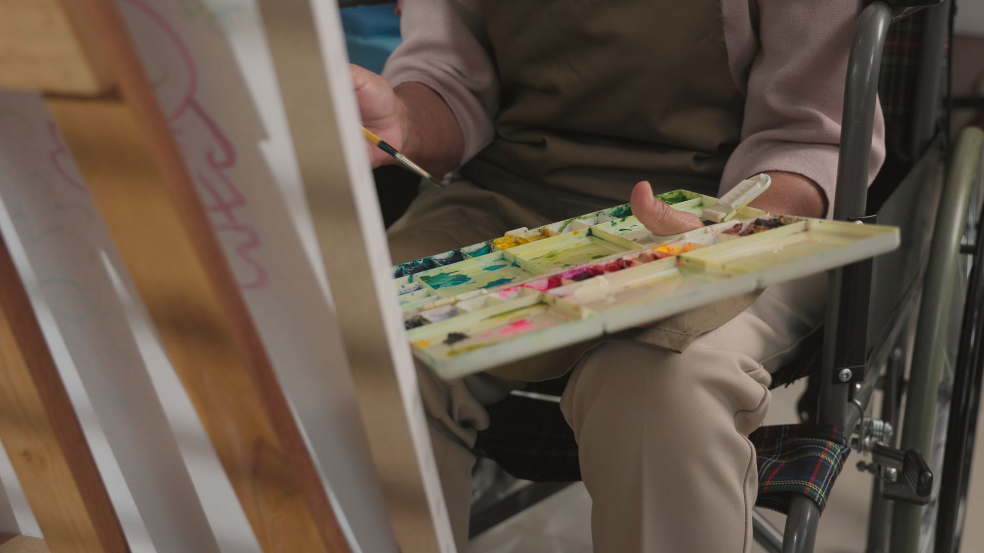 Person in wheelchair painting with palette and brush. Wooden easel and colorful paint visible.