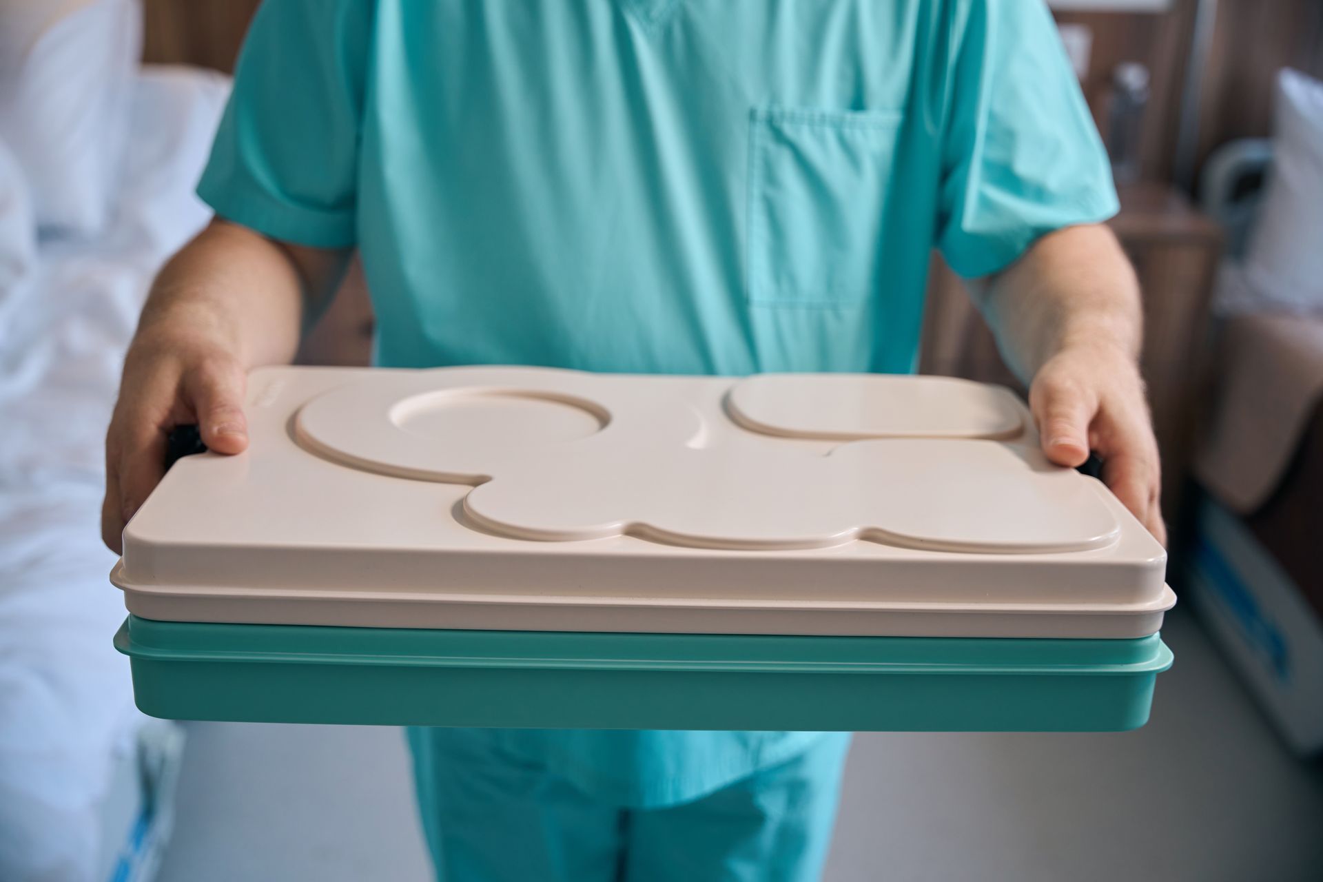 Person in teal scrubs holding a tray with beige and teal compartments, possibly for a meal, in a hospital room.