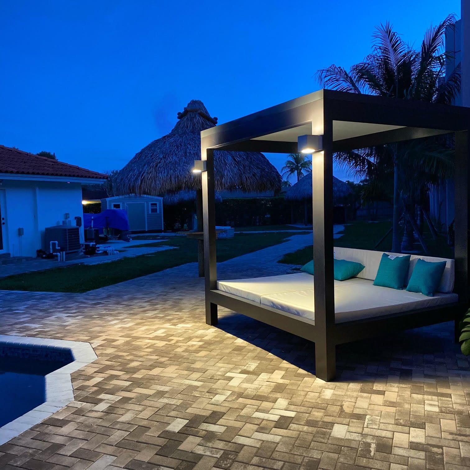 Outdoor lounge area with bed under a canopy, lit at dusk. Pool and thatched roof structure in the background.
