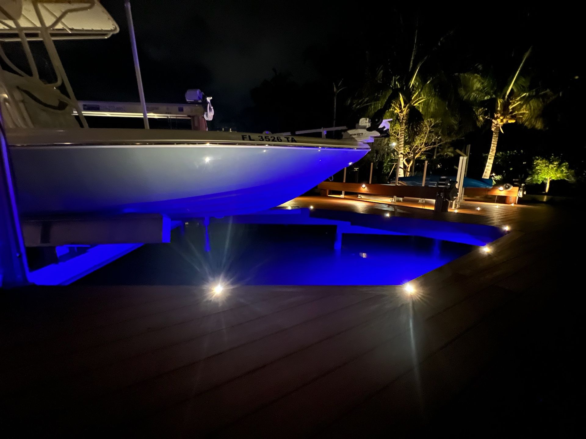 Boat on a dock at night, illuminated by blue and white lights. Palm trees and a dark pool in the background.
