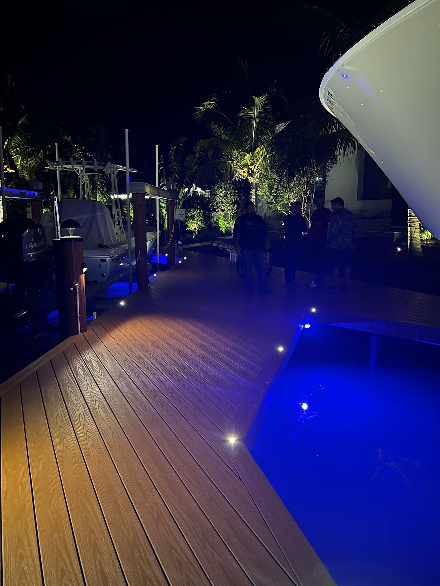 Wooden deck overlooking a pool lit with blue lights at night; people walking in the distance.