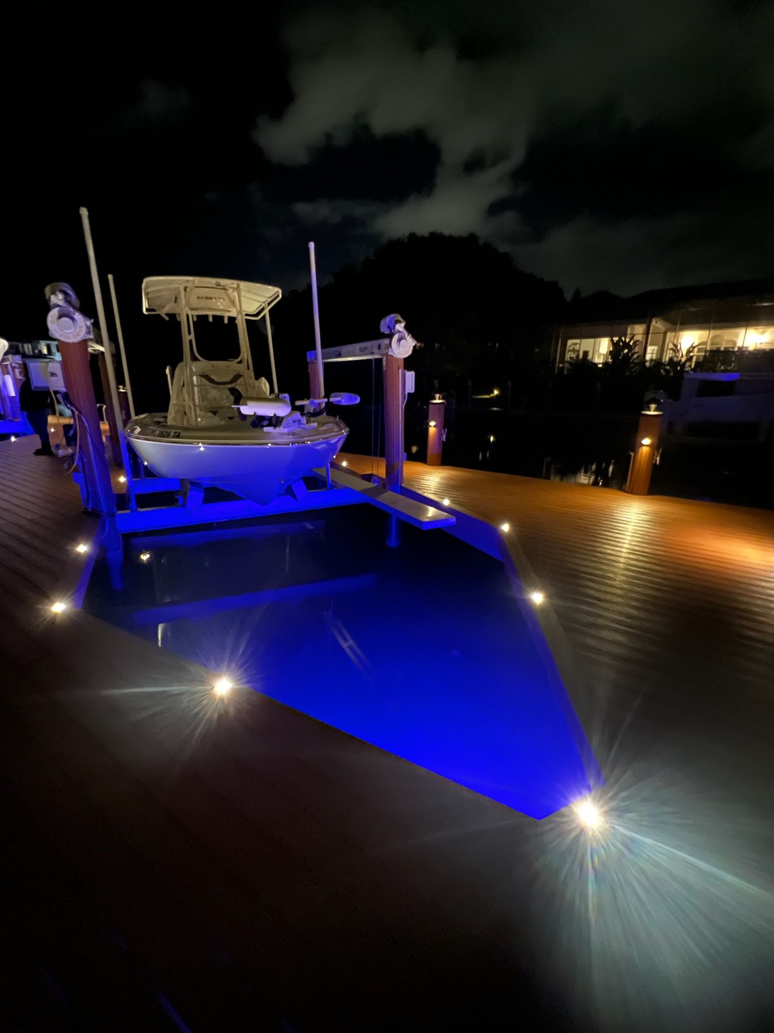 Boat on lift illuminated with blue light at night. Dock with lights and a gathering.