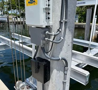Electrical boxes and conduits mounted on a concrete pole next to a boat lift, near water.