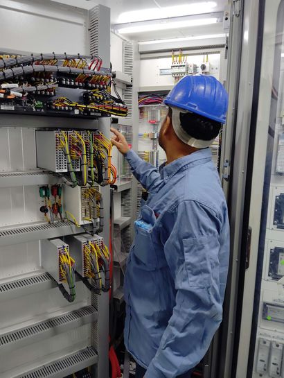 Electrician in a hard hat and gloves tests a fuse box with a multimeter.