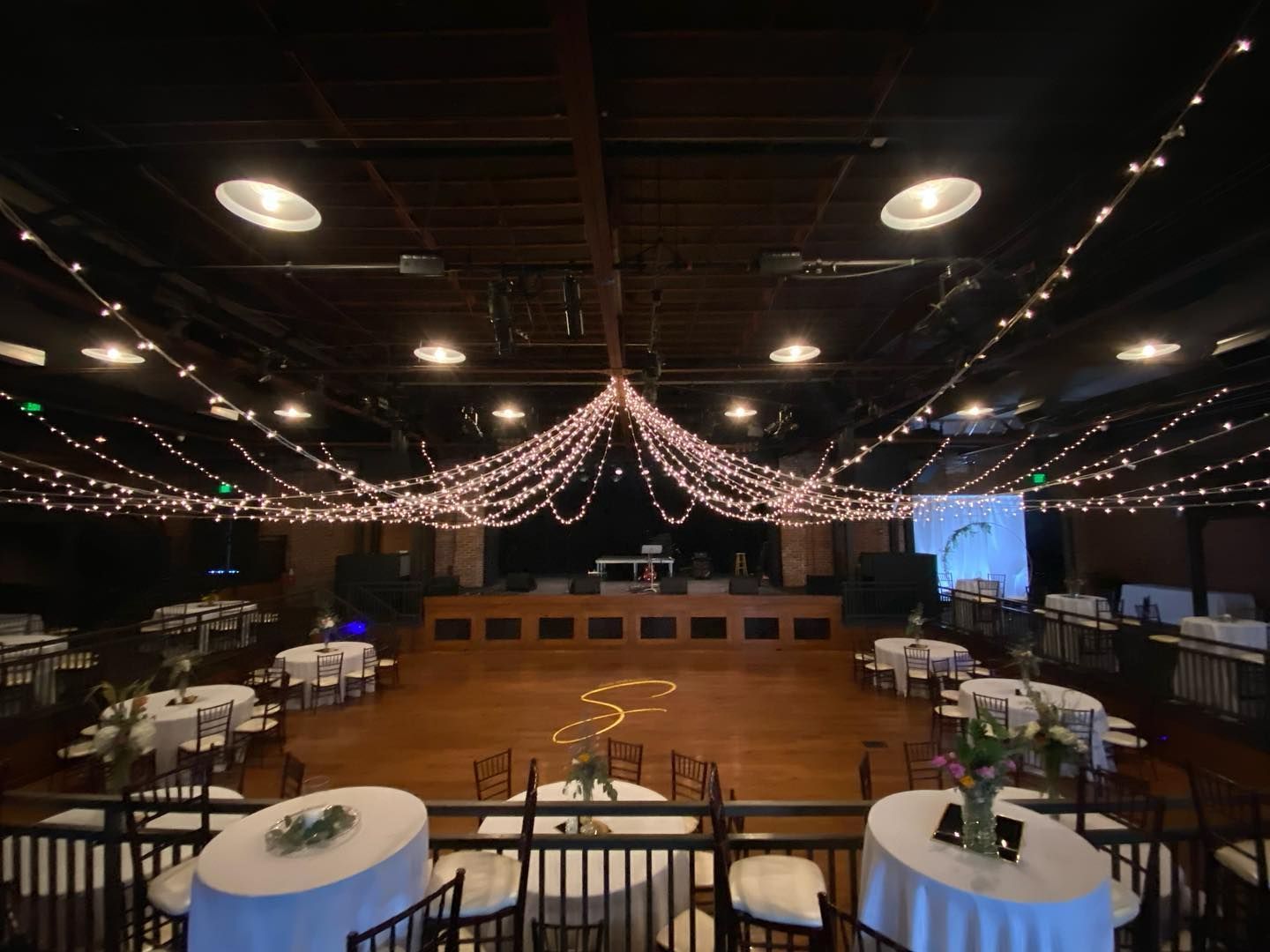 A large room with tables and chairs and a dance floor with lights hanging from the ceiling.