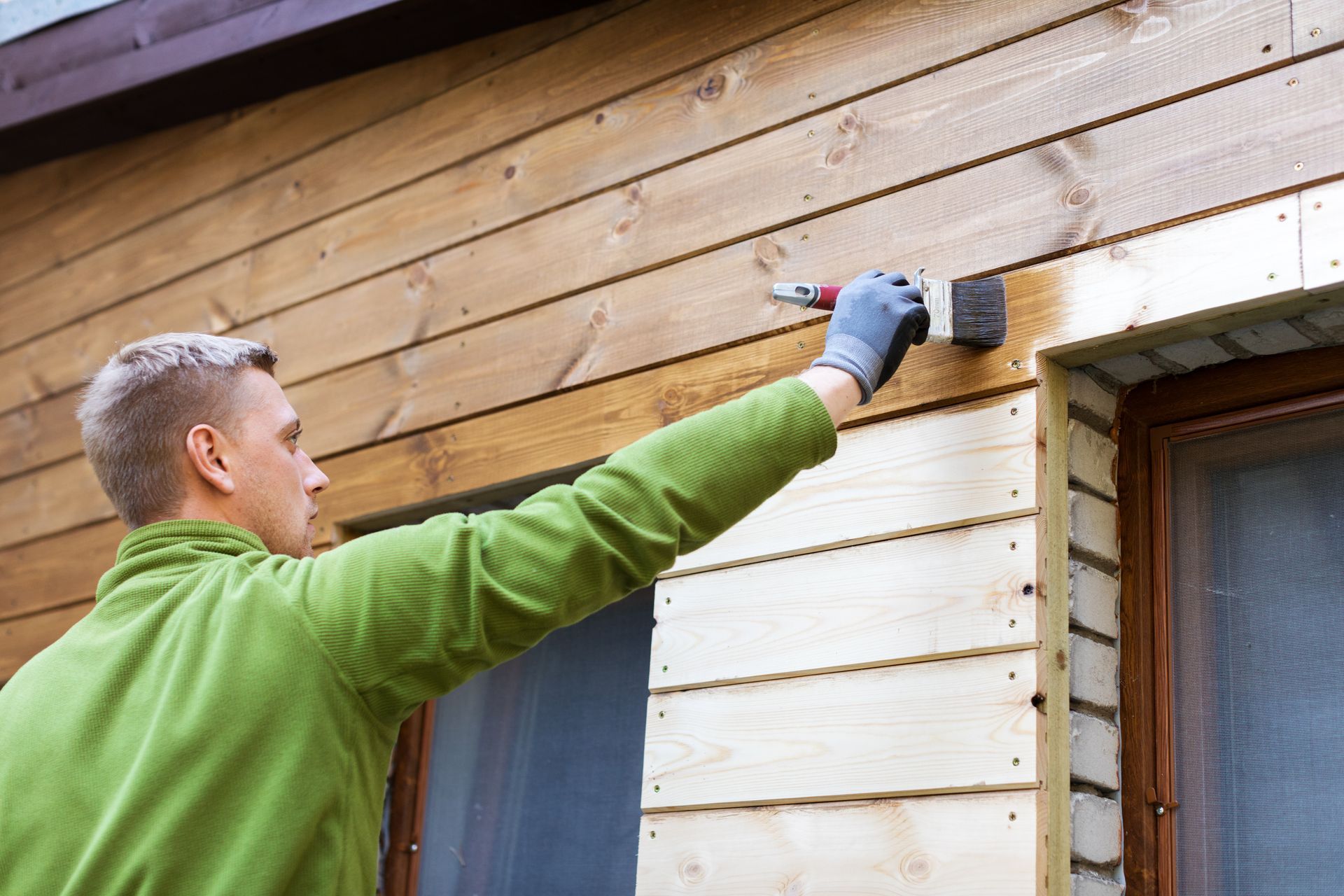 Man in green jacket staining wooden siding of a house with a brush.