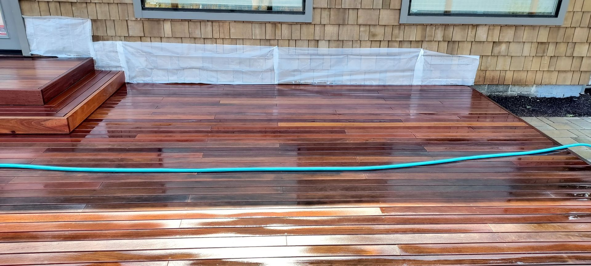 Wet wooden deck with a green hose, near a house with a white base.