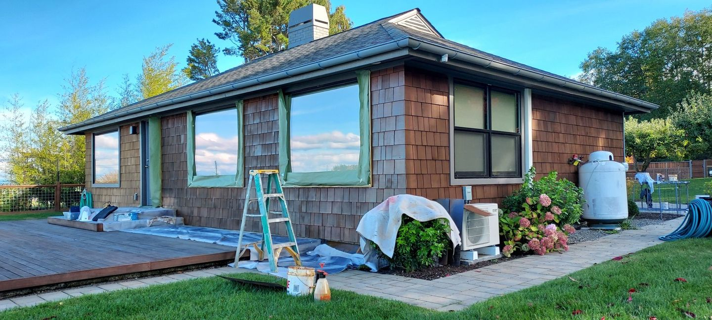 A house being renovated: brown shingles, large windows reflecting sky, ladder, painting supplies, and yard.