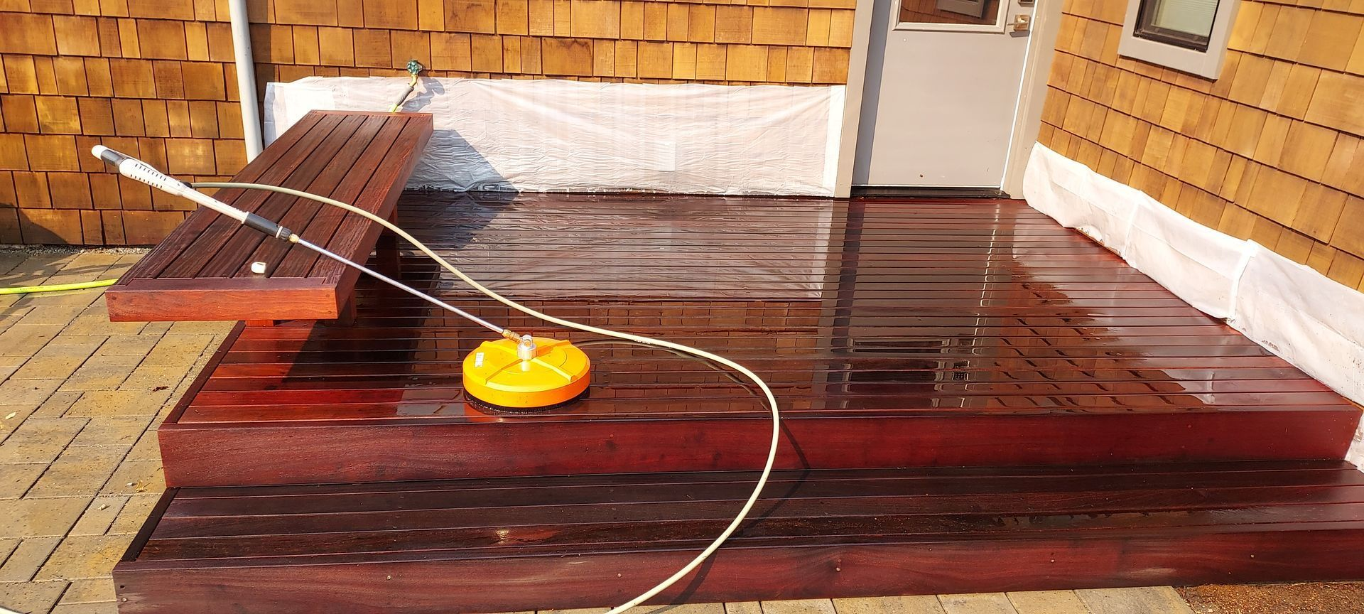 Wet wooden deck being cleaned with a pressure washer; yellow cleaning head on the deck.