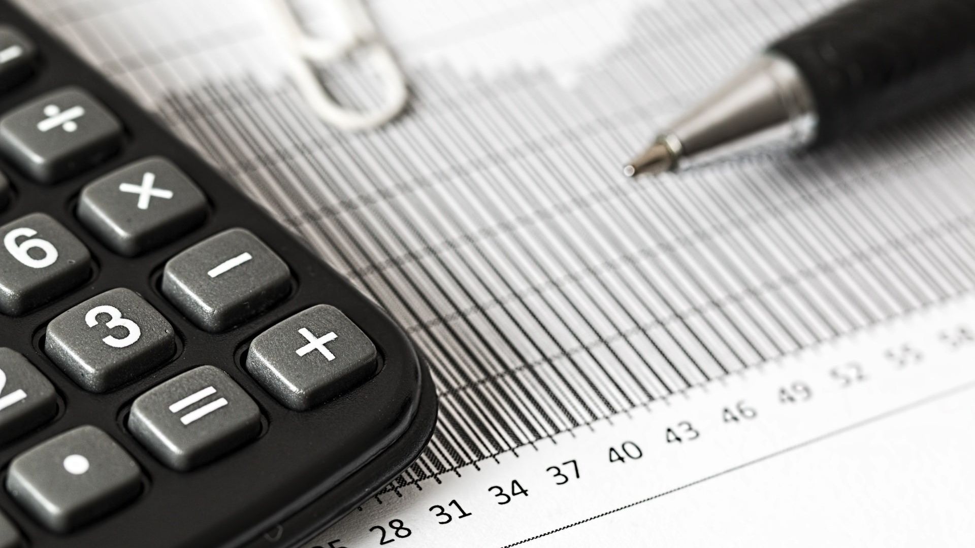 Close up photo of a desk with partial view of a calculator, pen, paperclip, and spreadsheet