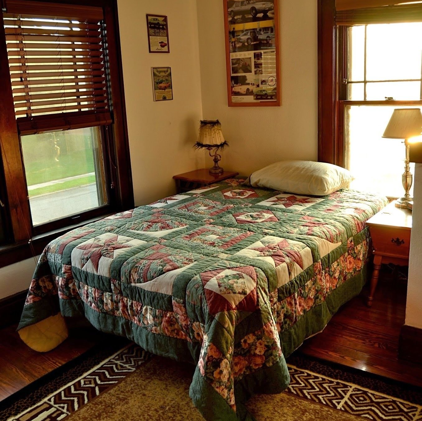Cozy bedroom with a floral quilt on a single bed, two windows with blinds, and a small nightstand.