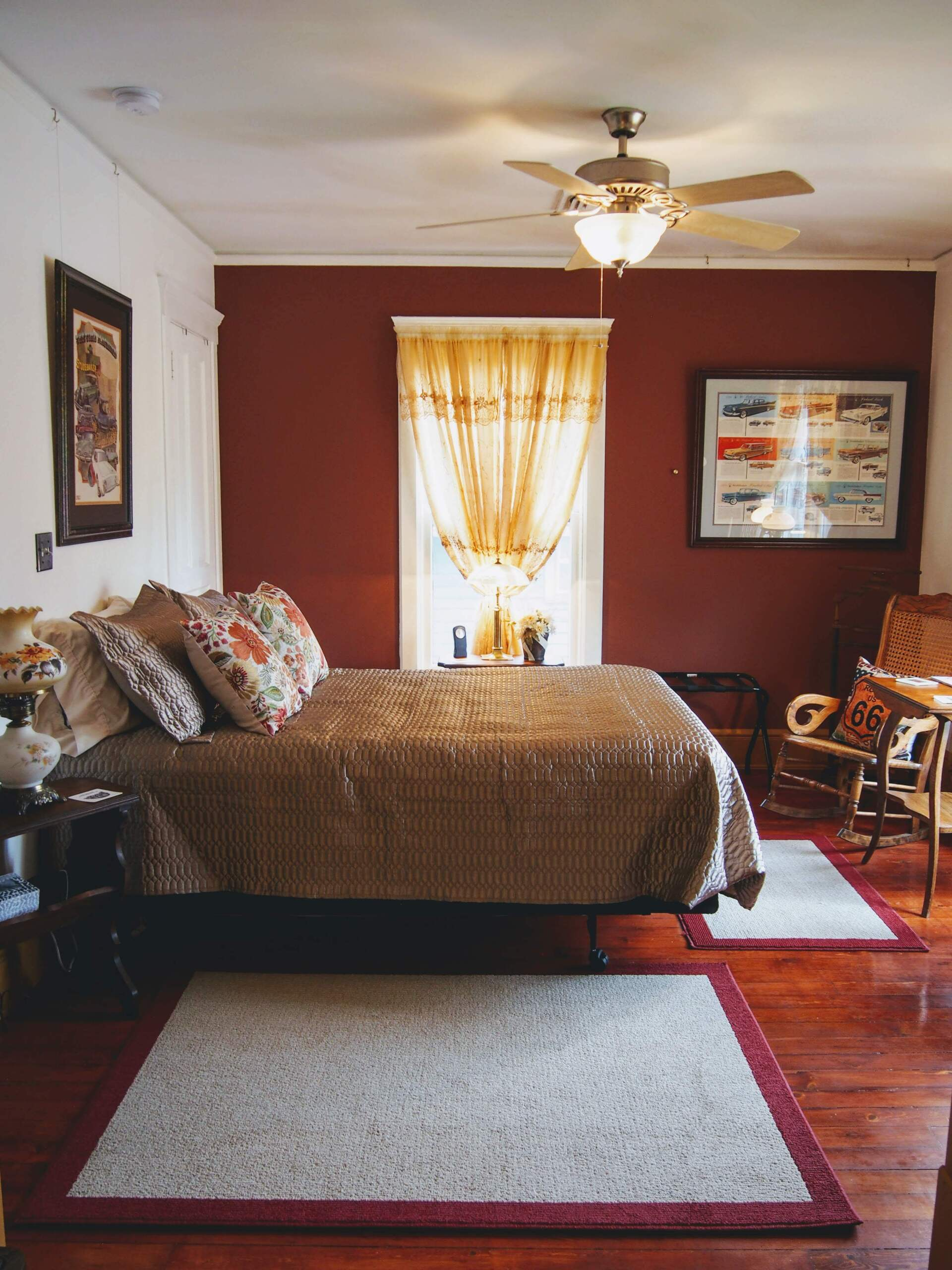 Bedroom with a red accent wall, bed with floral pillows, two area rugs, and wooden floor.