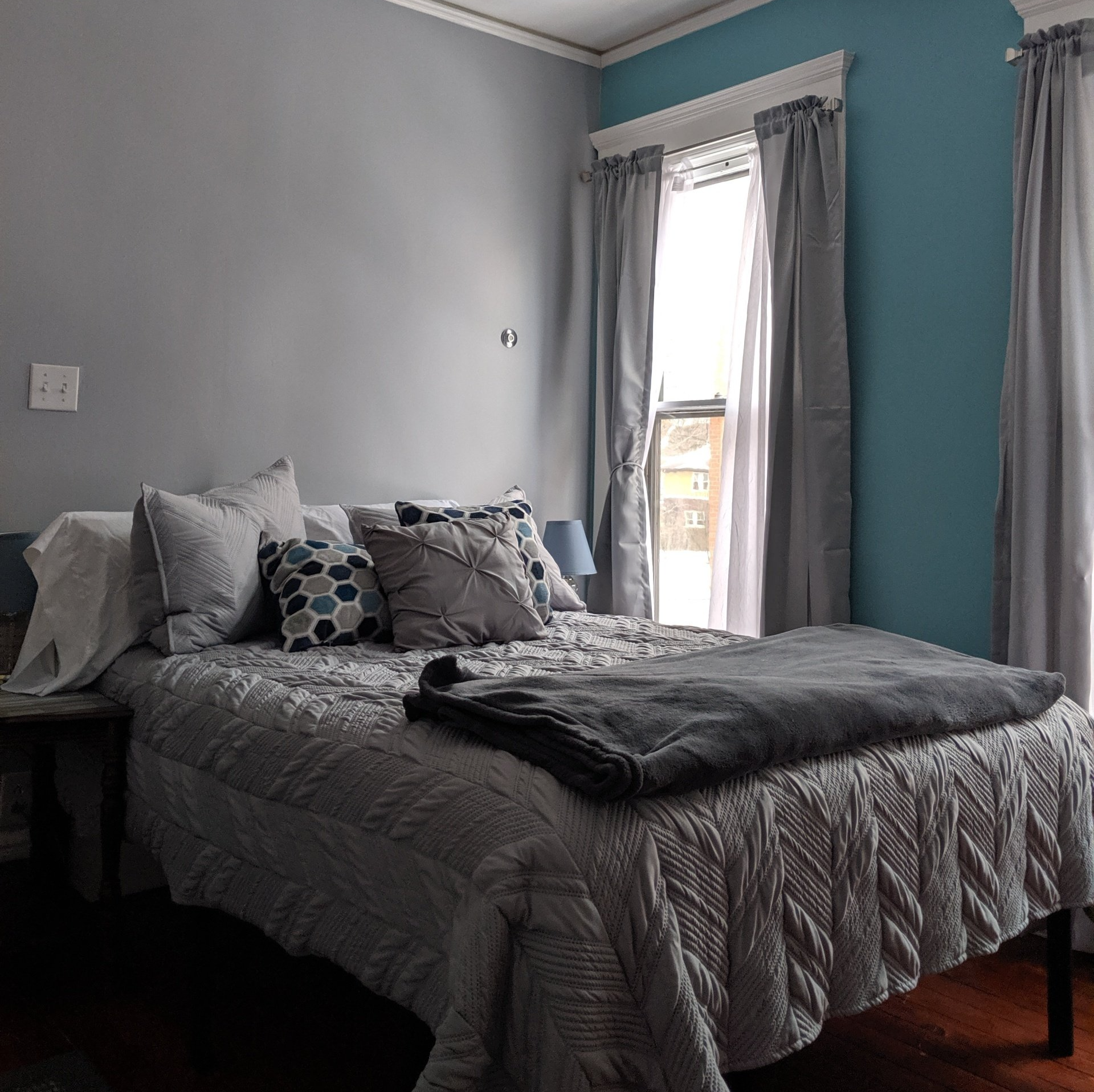 Bedroom with gray and teal walls, bed with gray comforter and pillows, window with gray curtains.