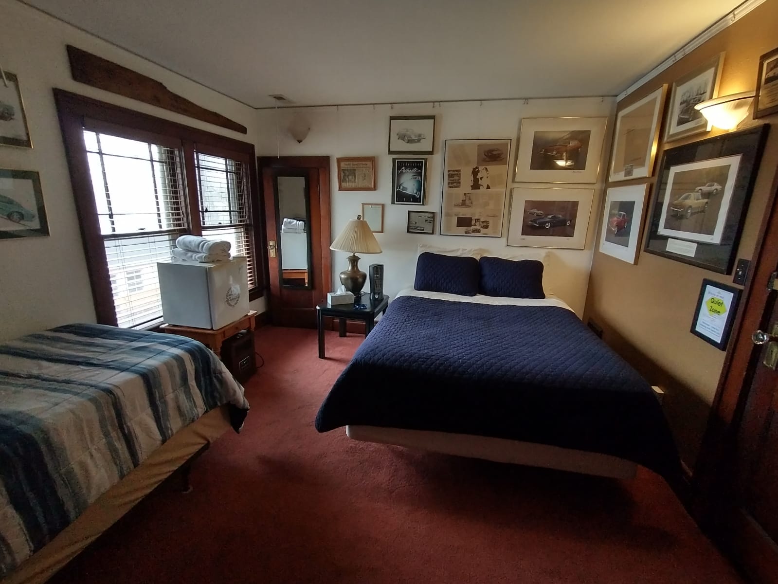 Bedroom with two beds, photos on walls, red carpet, dark blue bedspread, window, small table, lamp.