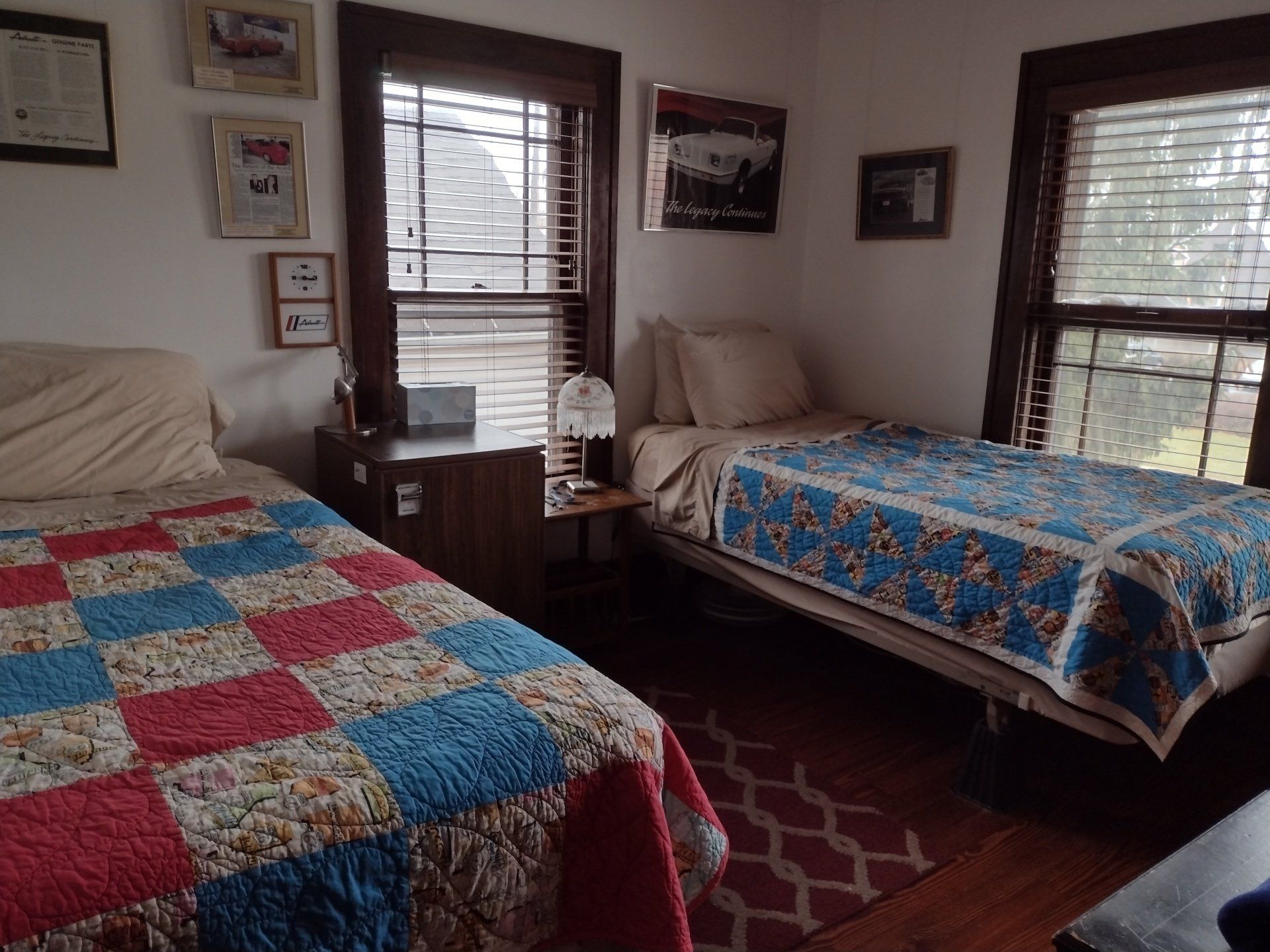 Two beds in a bedroom with quilts, two windows, and framed pictures on the wall.