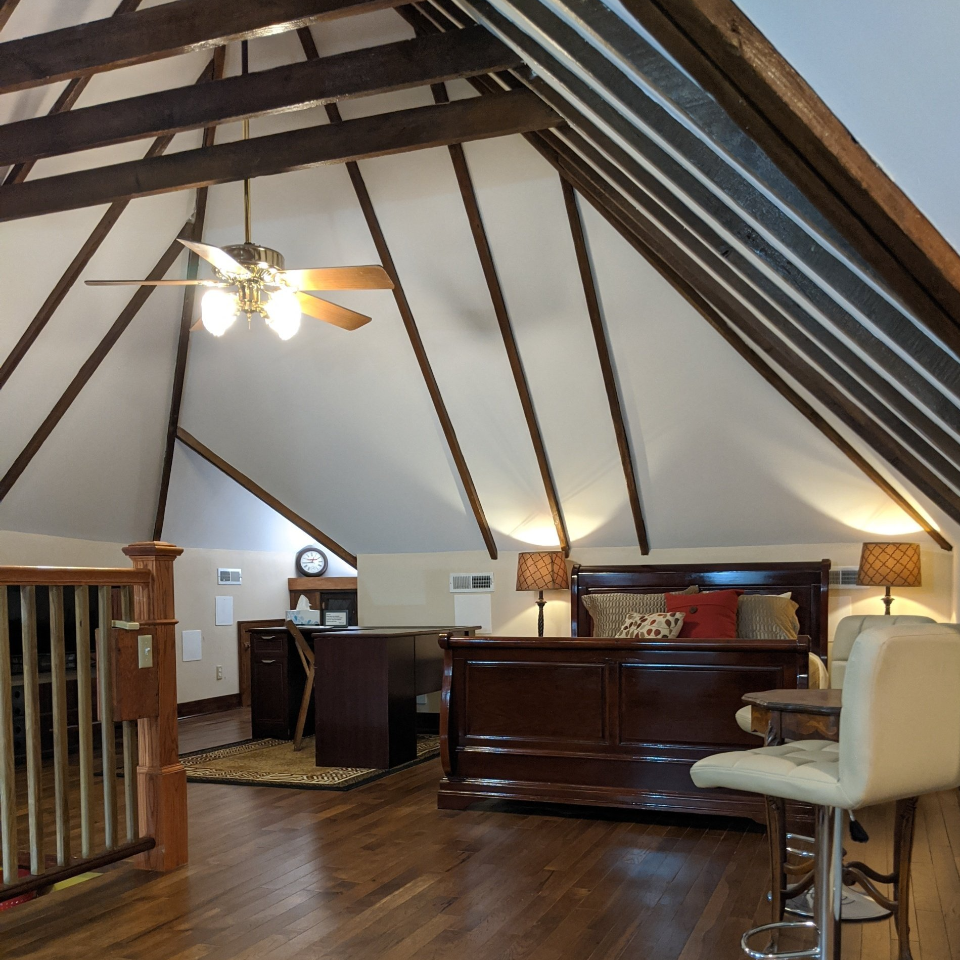 Bedroom with vaulted ceiling, dark wood furniture, desk, bed, and bar stools. Brown beams and ceiling fan.