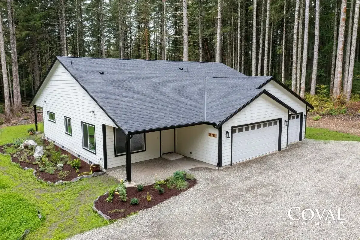 White house with dark gray roof, covered porch, and two-car garage in a wooded setting.